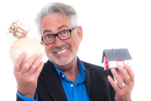 Ein älterer Mann hält in seinen Händen ein kleines Haus und ein gefülltes Sparschwein