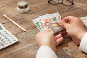 Ein Mann an einem Holztisch hält ein paar Geldscheine in der Hand. Ein Taschenrechner und ein Stift liegen auf dem Tisch
