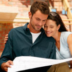 Junges fröhliches Paar steht vor einer Baustelle und studiert einen Bauplan